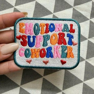 May include: A rectangular embroidered patch with a white background and a dark teal border. The text "EMOTIONAL SUPPORT COWORKER" is stitched in multi-colored letters including red, blue, pink, orange, and teal. Three small red hearts are below.