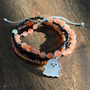 May include: A stack of three beaded bracelets featuring a white ghost charm with a heart-shaped mouth. The bracelets include translucent orange beads, small black seed beads, and iridescent metallic beads, all secured with a white adjustable cord.