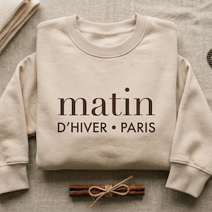 May include: A beige crewneck sweatshirt featuring the dark brown text "matin D'HIVER • PARIS" centered on the chest. The garment is displayed alongside a vintage-style pocket watch with a chain and a small bundle of cinnamon sticks tied with twine.