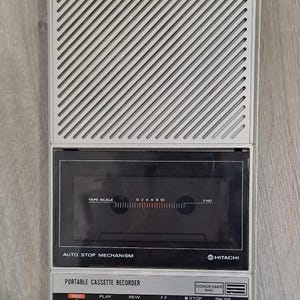 May include: A silver and black Hitachi portable cassette recorder featuring a large speaker grille at the top, a central tape compartment with a tape scale, and a control panel with buttons labeled REC, PLAY, REW, F.F, STOP, EJECT, and PAUSE.