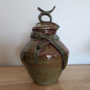 May include: A ceramic jar with a lid, featuring a glossy brown and green glaze. The jar has a rounded body and a decorative sculpted handle on the lid that resembles a crescent shape. A sculpted band wraps around the neck of the jar.