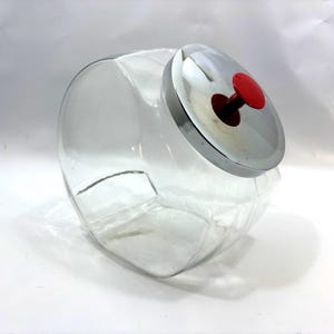 May include: A clear glass storage jar with a slanted opening and a circular silver metal lid. The lid features a small, bright red knob handle centered on top. The jar has a rounded, bulbous body shape and sits at an angle on its base.