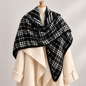 May include: A black and white plaid scarf draped over a cream-colored coat. The scarf features a classic checkered pattern with thin white lines intersecting on a black background, finished with a solid black border along the edges.