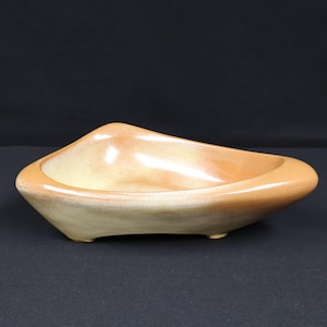 May include: A low-profile, triangular ceramic bowl with a smooth, glossy finish. The piece features a gradient of warm, earthy tones, transitioning from a light cream center to a deeper, golden-brown hue along the rounded, thick outer rim.