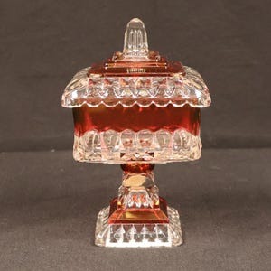 May include: A square glass candy dish with a lid, featuring a deep red color on the body and base, accented by clear, faceted, and pressed glass patterns. The piece stands on a pedestal base and includes a matching square lid with a finial top.