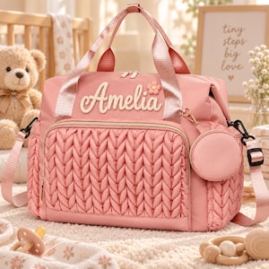 May include: A dusty pink diaper bag featuring a quilted cable knit front pocket, two top handles, and an adjustable shoulder strap. The name Amelia is embroidered in white script on the front, accompanied by a small flower and a round coin pouch.