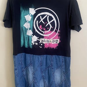 May include: A black short-sleeve t-shirt featuring a white Blink-182 smiley face logo, teal and pink brushstroke accents, and the text Blink-182. The lower portion of the garment is constructed from blue denim fabric with a diamond-stitched pattern.