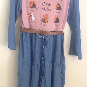 May include: A long-sleeve dress with a dusty pink bodice and blue sleeves and skirt. The bodice features a graphic of eight different chicken heads with the text "Comb Styles" and labels including "The Davy Crockett," "The Emo," and "The Trump." A brown belt with a double-ring buckle is worn at the waist.