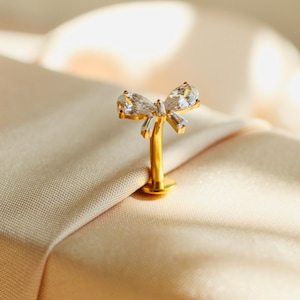 May include: A gold-toned body jewelry stud featuring a bow-shaped top adorned with two pear-cut clear stones and two baguette-cut clear stones. The piece includes a straight post and a flat circular base, designed for secure placement in a piercing.