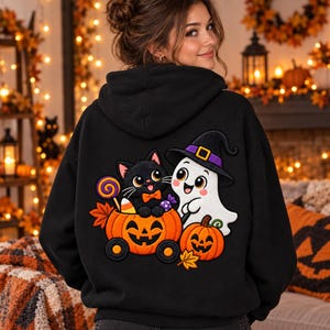 Puede incluir: Una sudadera negra con capucha que presenta un gran gráfico bordado en la espalda. El diseño muestra un gato negro con pajarita naranja sentado en un carro de calabaza, acompañado por un fantasma blanco con un sombrero de bruja morado.