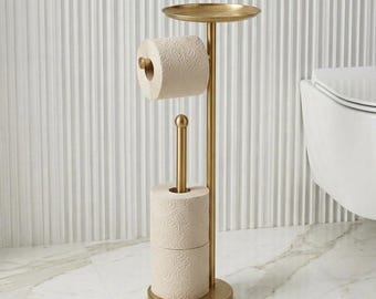 May include: A freestanding gold-toned metal toilet paper holder featuring a vertical pole with a top shelf, a horizontal arm for one roll, and a base post that holds two additional stacked rolls of textured cream toilet paper.