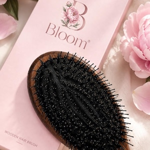 Könnte beinhalten: Eine Holzhaarbürste mit dunkelbraunem Griff und schwarzem Borstenkissen, die auf einer hellrosa rechteckigen Schachtel liegt. Die Schachtel zeigt den Text "Bloom" mit einer Blumengrafik und den Schriftzug "WOODEN HAIR BRUSH" in Schwarz.