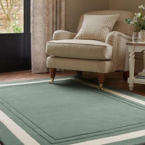 May include: A rectangular area rug featuring a solid sage green center field framed by a thick cream-colored border and a thin inner sage green line. The rug has a dense, low-pile texture and is positioned on a herringbone patterned wooden floor.