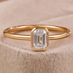 Könnte beinhalten: Ein goldener Ring mit einem einzelnen, im Smaragdschliff facettierten, klaren Edelstein in einer glatten Zargenfassung. Die Ringschiene ist schmal und poliert und weist eine reflektierende metallische Oberfläche auf. Der Stein ist rechteckig.