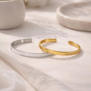 May include: Two open cuff bracelets, one in a polished silver tone and one in a polished gold tone. Both bracelets feature a smooth, flat, and minimalist band design with rounded ends, creating a simple and clean aesthetic for wrist wear.