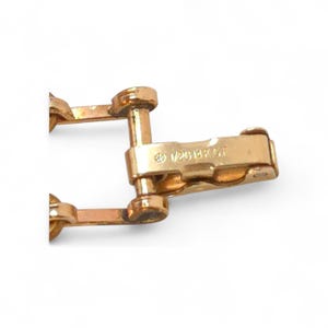 May include: Close-up of a gold-toned jewelry clasp. The clasp has a rectangular shape with rounded edges and a hinged mechanism. The text "1/20 14K G.F." is stamped on the clasp, indicating the material composition. The jewelry clasp is a component of a bracelet or necklace.