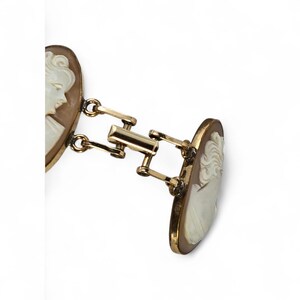 May include: A pair of gold-toned cufflinks featuring oval cameo portraits. The cameos are light brown with white relief carvings of a classical figure. The cufflinks are connected by a series of gold-toned bars and hinges.