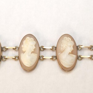 May include: A gold-toned bracelet featuring oval cameo pendants. Each cameo displays a profile of a woman in relief, set against a light peach background. The bracelet links are connected by small gold bars.