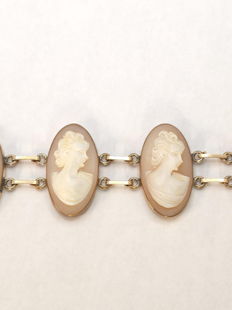 May include: A gold-toned bracelet featuring oval cameo pendants. Each cameo displays a profile of a woman in relief, set against a light peach background. The bracelet links are connected by small gold bars.