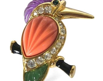 May include: A gold-tone bird brooch featuring a purple carved crest, a large coral-colored teardrop-shaped body, and a green carved tail. The bird is perched on a black branch, accented with clear rhinestones along the body and a small green eye.