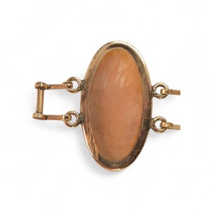 May include: A gold-toned oval cameo bracelet clasp. The clasp features a light brown oval cameo set within a gold frame. The clasp has a rectangular bar and two sets of loops for attachment.