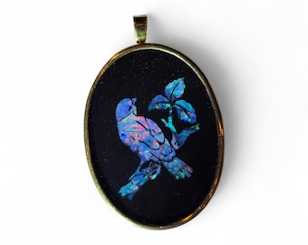 May include: Oval pendant with a gold-tone frame. The pendant features a bird perched on a branch with leaves, crafted from iridescent blue and pink materials against a black background. The pendant has a small loop at the top.