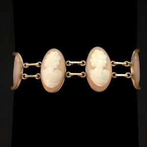 May include: A gold-toned bracelet featuring three oval cameo pendants. Each cameo depicts a profile of a woman with detailed hair and clothing. The bracelet has a classic design, with connecting links and curved end pieces.