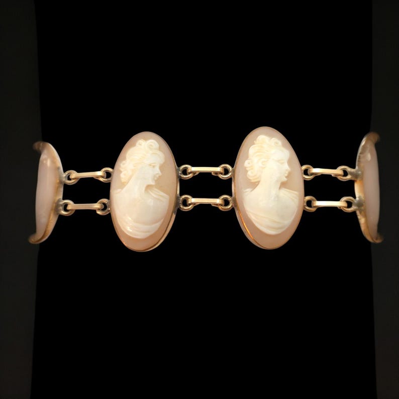 May include: A gold-toned bracelet featuring three oval cameo pendants. Each cameo depicts a profile of a woman with detailed hair and clothing. The bracelet has a classic design, with connecting links and curved end pieces.