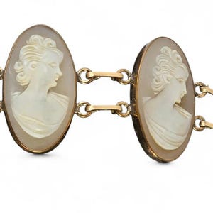 May include: A gold-toned bracelet featuring two oval cameo pendants. Each cameo displays a profile of a woman with detailed hair and draped clothing. The cameos are set in gold frames and connected by delicate gold links.