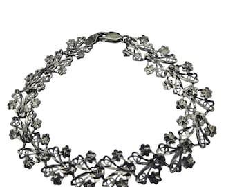 May include: A silver-toned filigree bracelet featuring a delicate floral design. The bracelet is composed of interconnected floral and leaf-like shapes, creating an openwork pattern. It has a secure clasp closure and is suitable for various occasions.