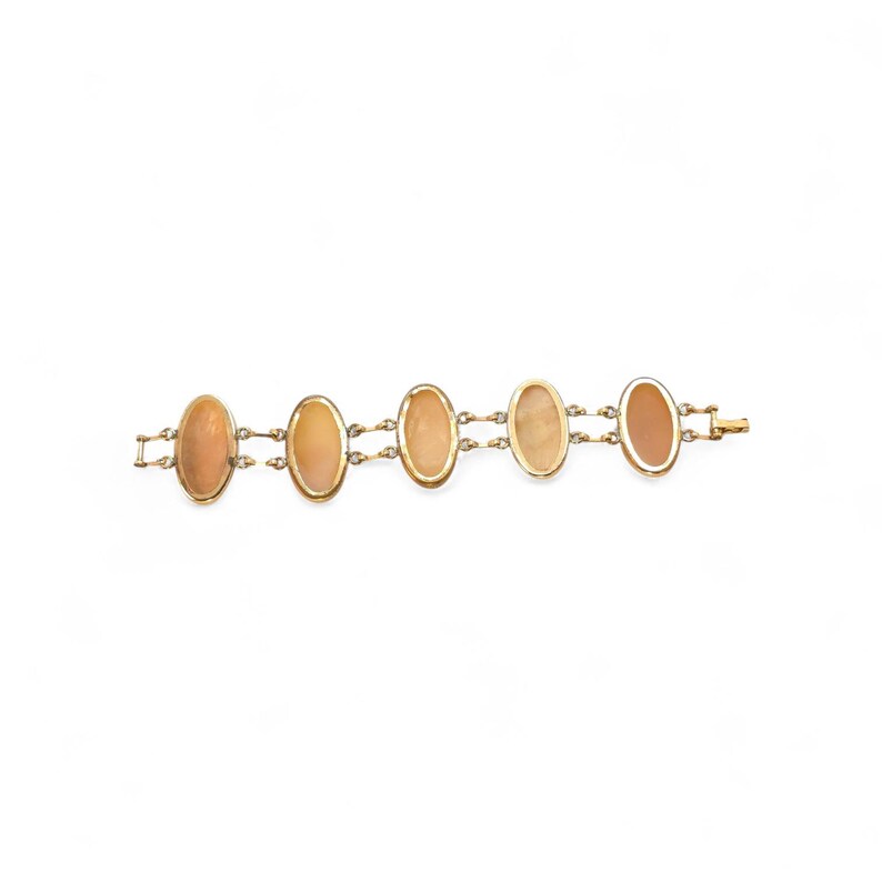 May include: A gold-toned bracelet featuring five oval-shaped, peach-colored stones. The stones are linked together with small gold chains and set in gold frames. The bracelet has a simple clasp closure.