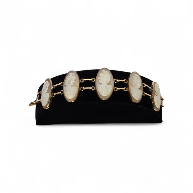 May include: A gold-toned bracelet featuring oval cameo pendants with white relief portraits. The bracelet is displayed on a black velvet surface. The cameo pendants are connected by small gold-toned links.