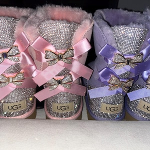 May include: Two pairs of boots, one pink and one purple, featuring dense rhinestone embellishments on the shafts and heels. Each boot is adorned with two satin bows and metallic rhinestone-encrusted bow accents. The UGG logo is visible on the heel.