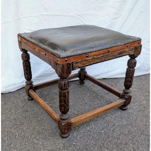 May include: A square footstool featuring a dark brown leather top secured with metal studs. The base is constructed from dark stained wood with four carved legs connected by horizontal stretchers. A light brown textured trim borders the seat.