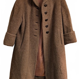 May include: A brown textured coat with a fuzzy, mohair-like finish, featuring a high collar, long sleeves with folded cuffs, and a front closure with five dark brown circular buttons. The interior is lined with a smooth, light brown fabric.