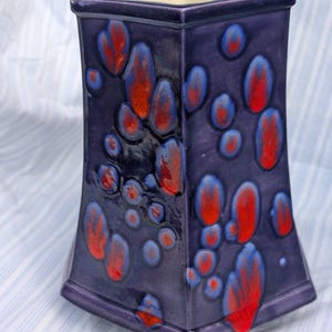 May include: A hexagonal ceramic vase with a glossy deep purple glaze featuring a crystalline pattern of vibrant red and blue circular spots. The interior of the vase is a smooth, off-white colour, and the exterior has a flared base and rim.