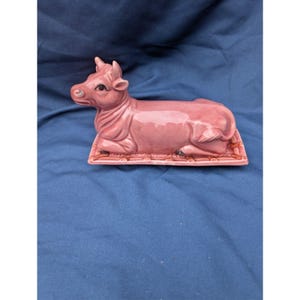 May include: A glossy pink ceramic butter dish shaped like a reclining cow. The cow figure rests on a rectangular base with a textured, grid-like pattern. The piece features a smooth, glazed finish with defined facial features and ears.