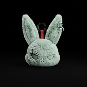 May include: A plush rabbit head keychain in a soft sage green color. The rabbit features two long ears, one with two silver metal rings, and a red fabric loop with a black ring. The face has stitched scars, closed eyes, and a small white nose.
