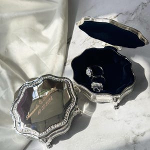 Könnte beinhalten: Eine silberfarbene Schmuckschatulle mit gewelltem Rand und Klappdeckel. Der Deckel ist mit dem Text "Amanda & Anthony 10.07.2028" graviert. Das Innere ist mit dunkelblauem Samt ausgekleidet und enthält zwei silberne Ringe mit klaren Steinen.