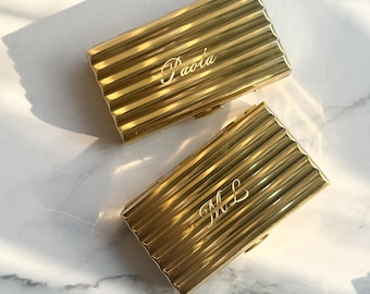 以下が含まれることがあります： リブ状の波打つ表面テクスチャを持つ、2つの長方形のゴールドトーンの金属製ケース。一方のケースには筆記体で「Paola」という名前が刻印されており、もう一方のケースには同じ筆記体スタイルで「ML」というイニシャルが刻印されています。