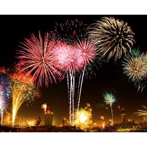 May include: A vibrant display of fireworks illuminating the night sky with bursts of red, pink, gold, and blue light. The explosions create starburst patterns and trailing sparks above a dark, silhouetted horizon line at night.