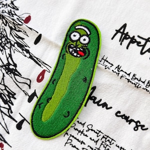 May include: An embroidered patch shaped like a green pickle character with large white eyes, a wide open mouth, and a red tongue. The character has a light green center and a dark green border, featuring a cartoon-like expression with black outlines.