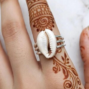 Puede incluir: Un anillo de plata con una concha de cauri blanca como punto focal central. La banda del anillo está construida con tres hilos de plata finos y paralelos que se conectan a la concha, creando un diseño abierto y en capas en el dedo.