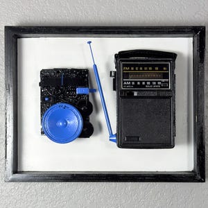 May include: A black framed wall art piece featuring a disassembled black radio with a prominent blue speaker and a blue antenna. The radio components are mounted on a white background, displaying AM and FM frequency scales and solid state text.