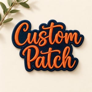May include: An embroidered patch featuring the text Custom Patch in bright orange cursive lettering against a dark navy blue background with a thick navy blue border that follows the contour of the words.