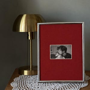 May include: A rectangular picture frame with a thin silver metallic border and a deep red matting. Centered within the matting is a small black and white photograph depicting a person kissing a cat. The frame stands upright on a flat surface.