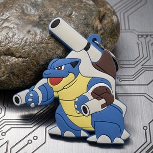 May include: A rubberized keychain featuring a blue, yellow, and white creature with two large cannons mounted on its back and smaller cannons on its arms. The character has a brown shell, a red mouth, and a metal ring attached to the top for keys.
