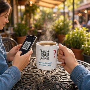 May include: A white ceramic mug featuring a printed QR code, a red question mark, and the text "Personalise your message Here" in blue. The mug contains a steaming hot dark beverage and is held by a hand near a smartphone.