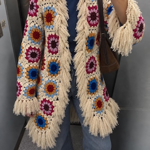 May include: A long-sleeved crochet cardigan constructed from multi-colored granny squares in shades of cream, bright pink, deep red, and royal blue. The garment features long cream-colored fringe trim along the front opening and sleeve cuffs.