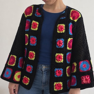 Könnte beinhalten: Eine schwarze, vorne offene Strickjacke aus gehäkelten Granny Squares. Die Quadrate zeigen leuchtende kreisförmige und florale Muster in Neonpink, leuchtendem Gelb und Elektroblau. Das Kleidungsstück hat lange, weite Ärmel und einen geraden Saum.